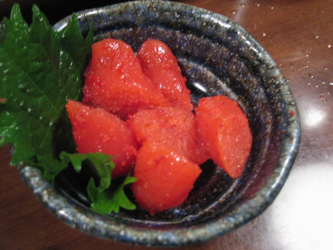 Slices of bright red mentaiko, cured pollock roe, in a dark bowl with green herb leaves