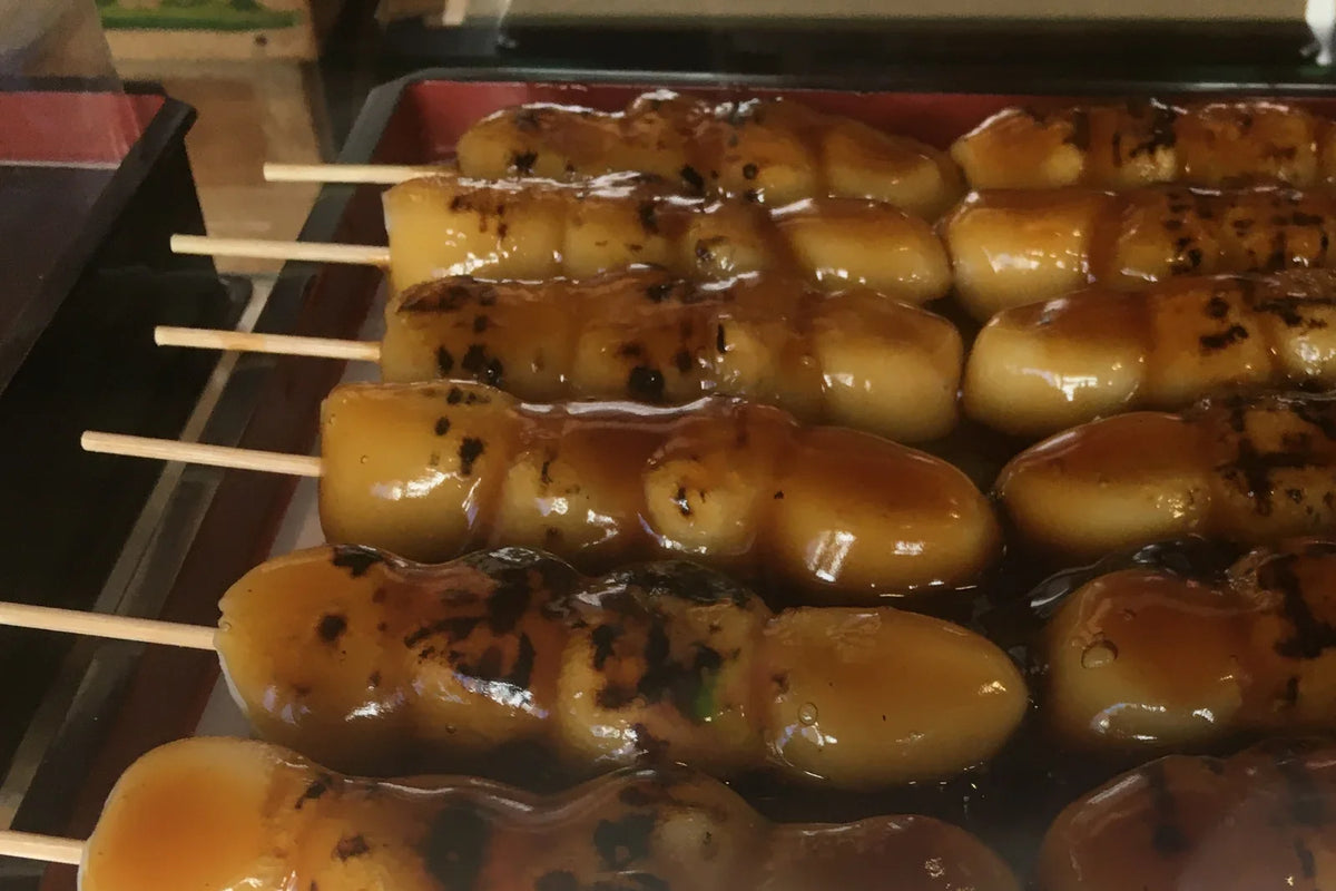 Rows of dango on bamboo skewers under a glossy brown glaze, on a tray at a Japanese stall