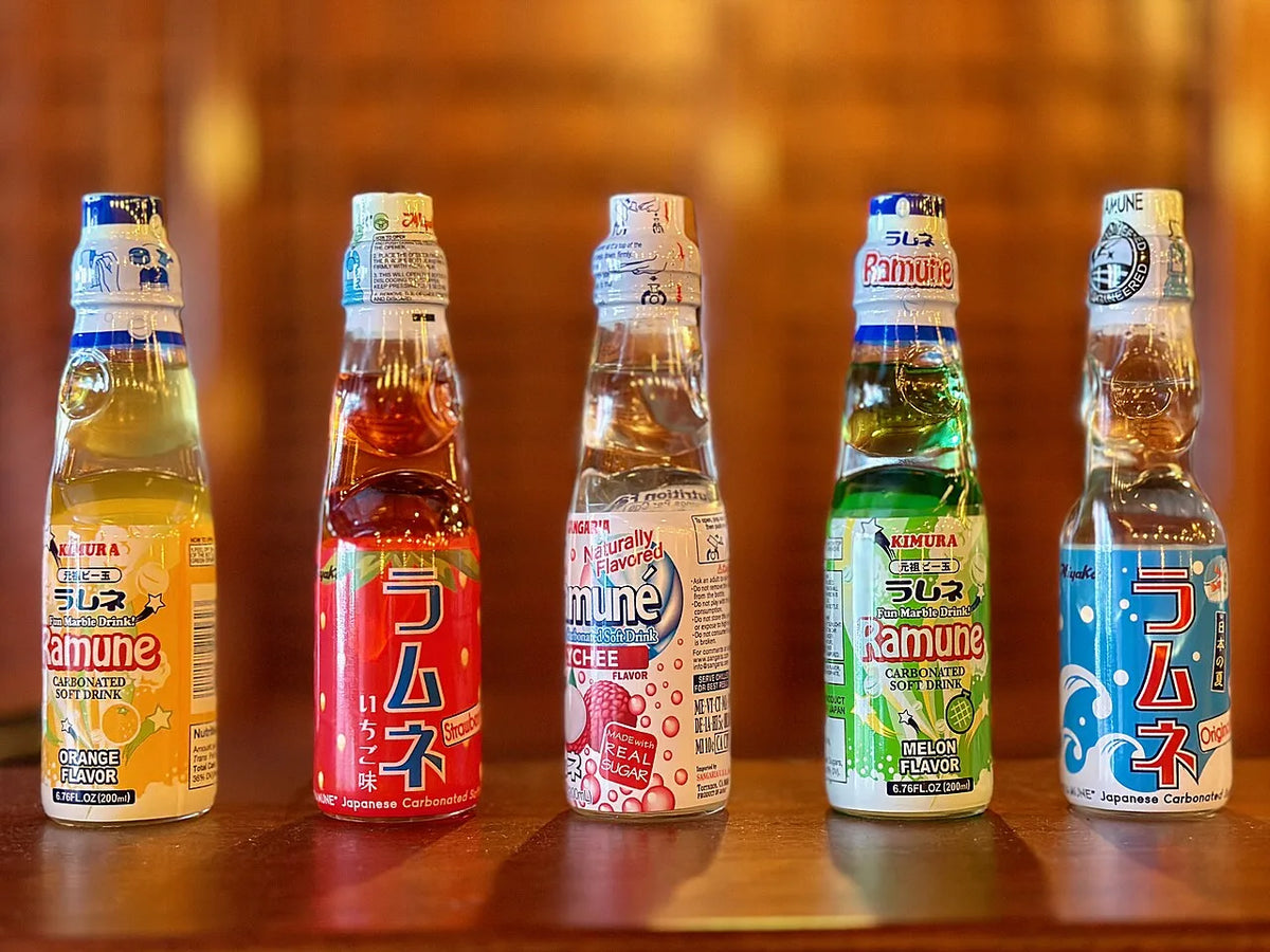 Five bottles of ramune in a row — orange, strawberry, lychee, melon and original — each with a glass marble in its neck