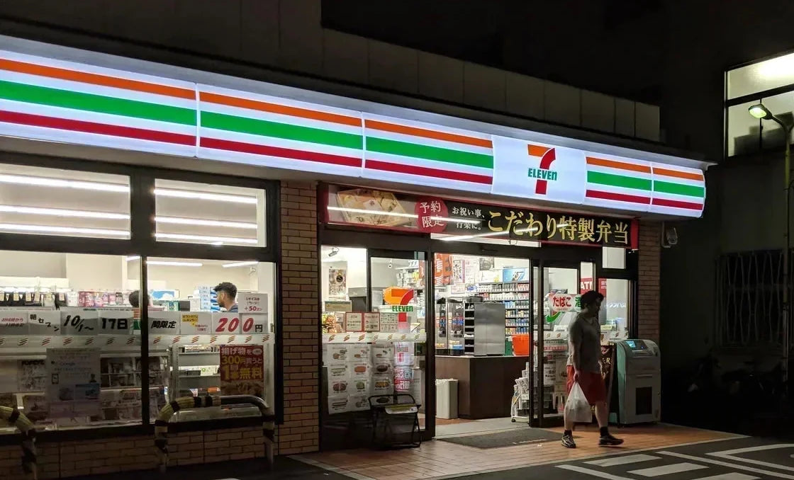A 7-Eleven in Japan at night, a customer stepping out through the lit doorway